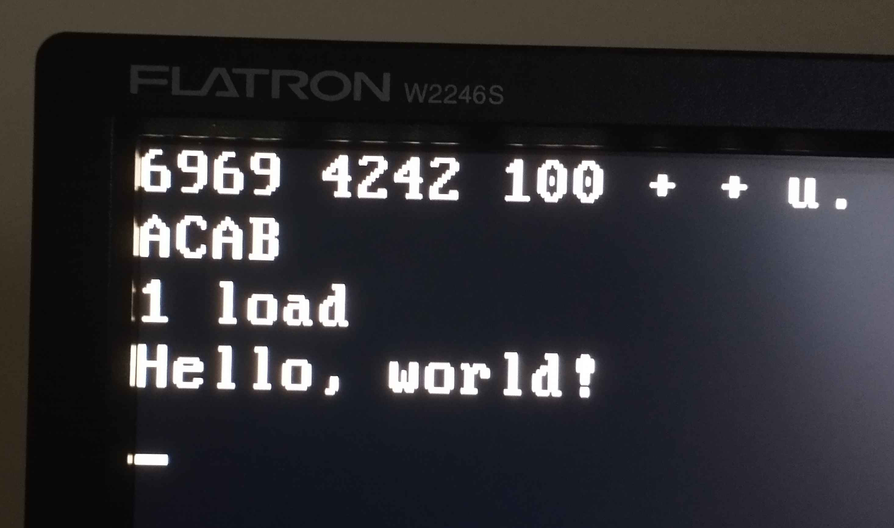 A photo of the corner of a monitor. We see the calculation "6969 4242 100 + + u-dot", which results in "ACAB". Then the "1 load" command is issued, to which the system describes "Hello, world!"